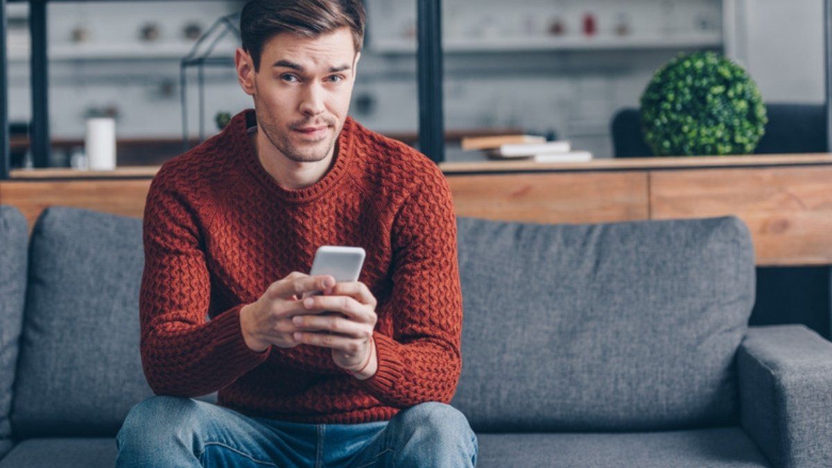 Man in a red jumper looking doubtfully at a suspicious message on his phone, spotting new scams