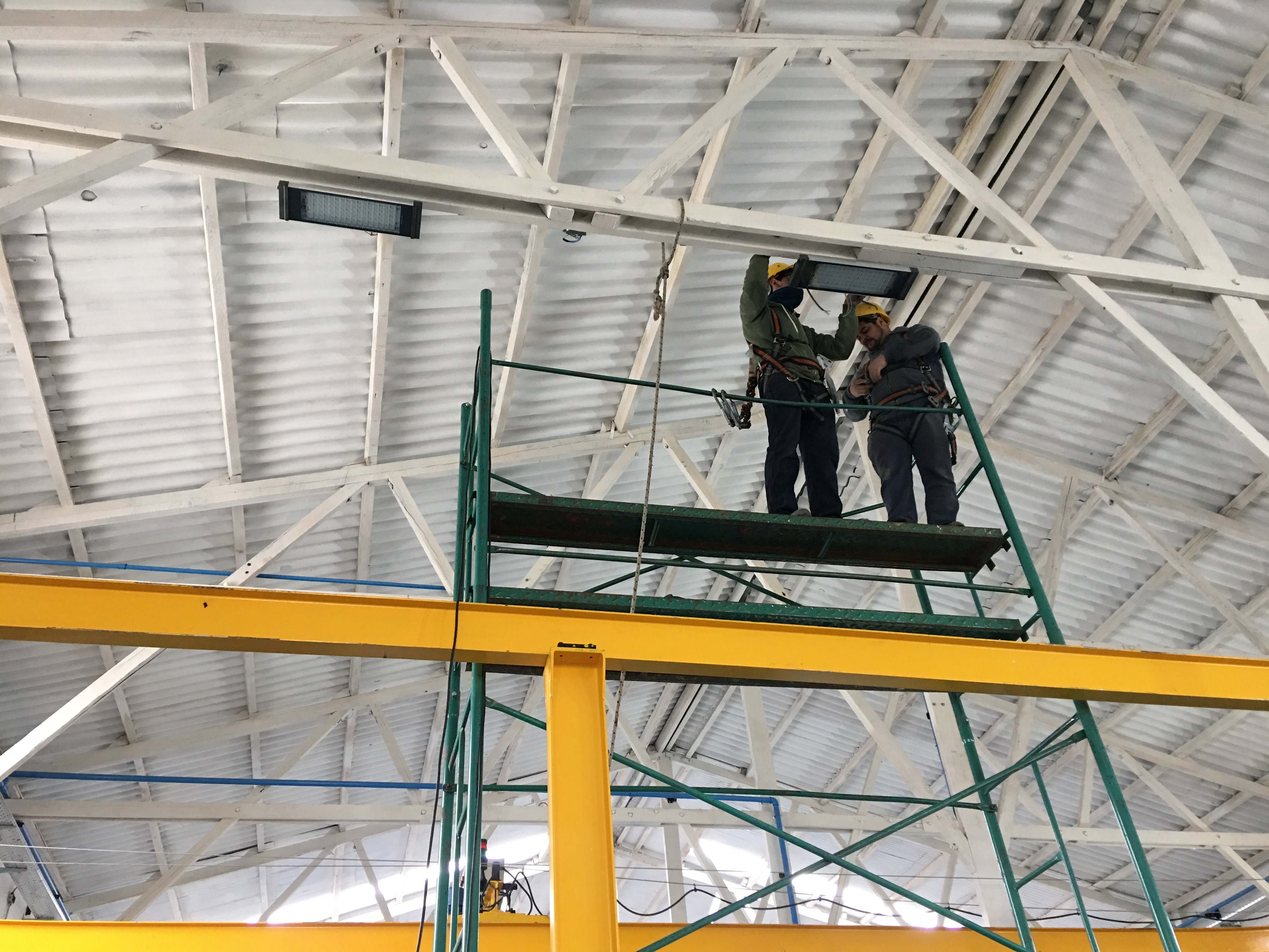 Dos operarios con arneses instalan lámparas LED en el techo de una nave industrial.