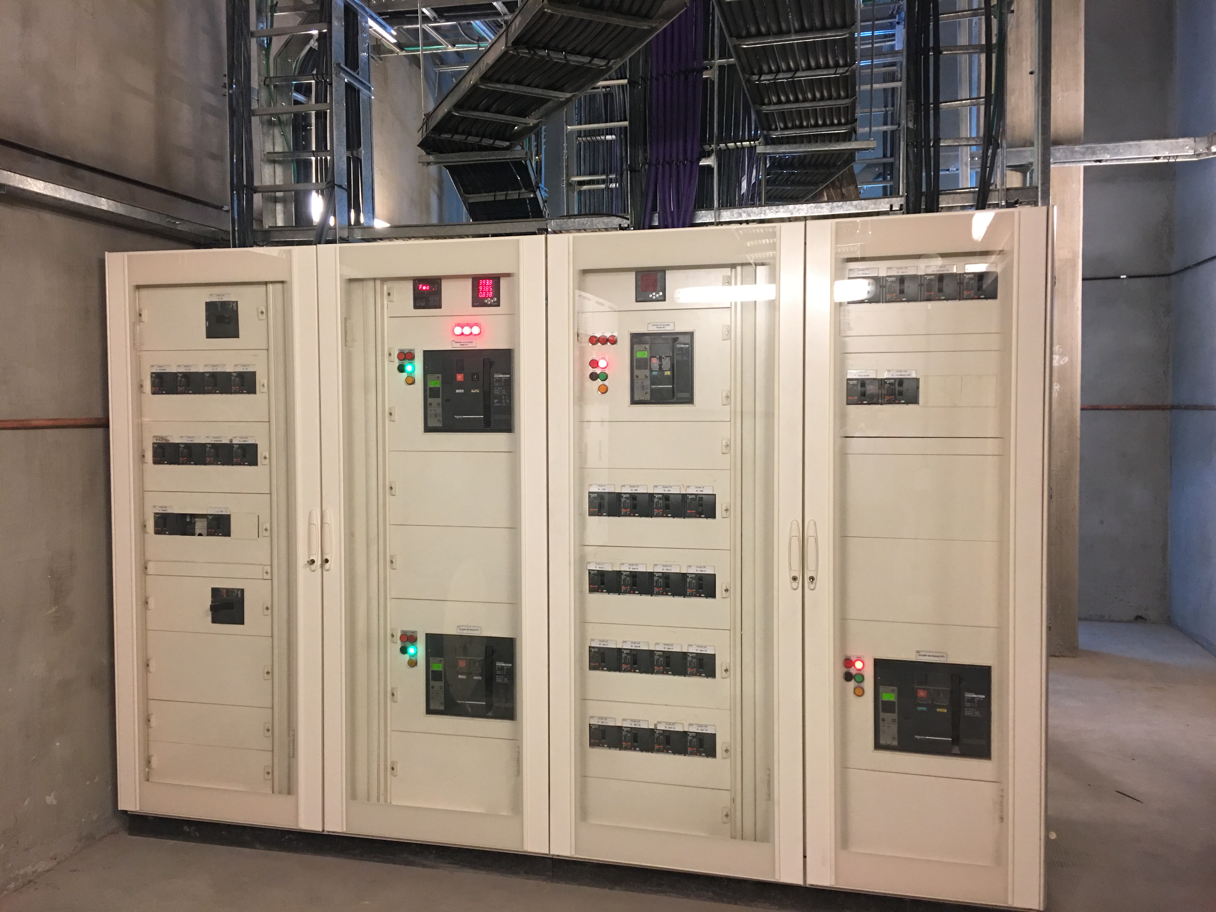 Panel eléctrico industrial con interruptores y luces indicadoras bajo bandejas de cables en sala técnica.