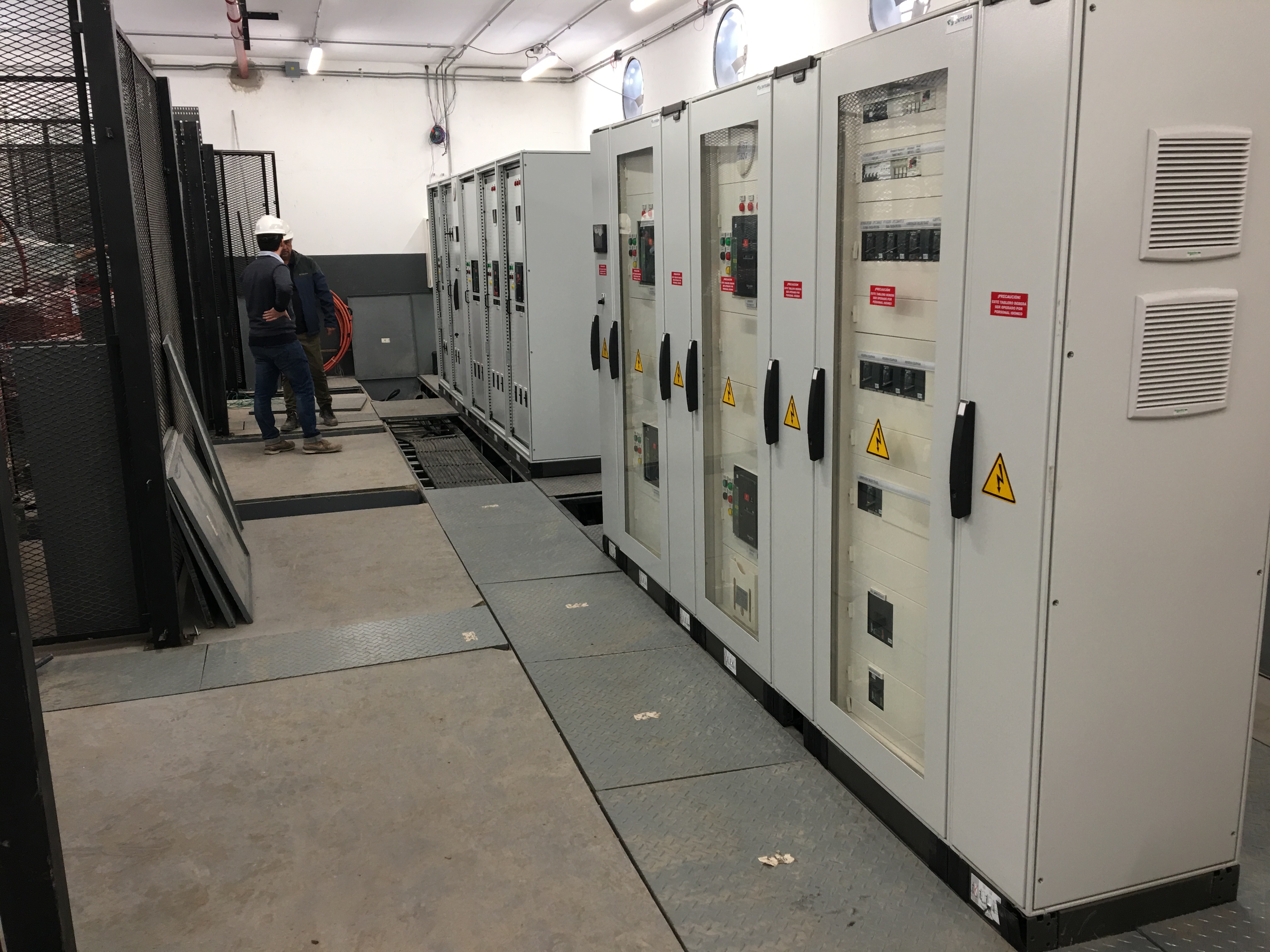 Paneles de control eléctrico industriales con señales de advertencia y dos técnicos en una sala.